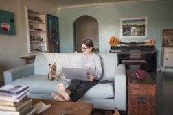 一个女人戴着眼镜坐在蓝色的沙发上跟她回家的创意笔记本电脑和小的狗”width=