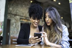 两个女人坐在咖啡馆里，看着智能手机“width=