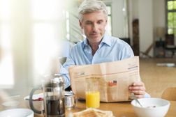 年长的男人吃早餐和阅读纸张“width=