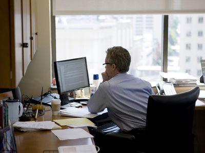 坐在有膝上型计算机的一张书桌的一个体贴的人的背面图在显示一个城市地平线的窗口前面