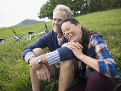 一对笑着的退休夫妇坐在两辆自行车旁边的草地上。