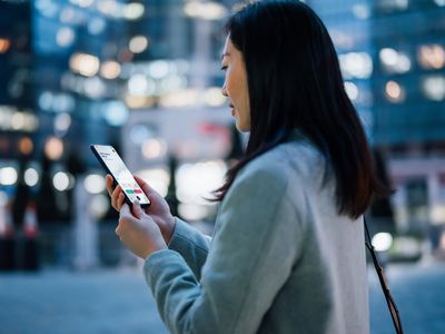 检查与在智能手机的移动应用程序的年轻亚裔女实业家财政股市分析在去的智能手机，反对在金融区的被照明的当代公司摩天大楼在晚上。