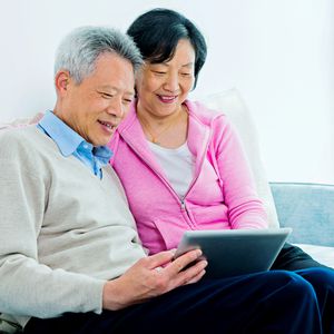 微笑的退休夫妇一起坐在沙发上看平板电脑”width=