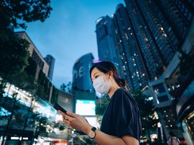 有保护的面罩的年轻亚裔妇女使用在去的智能手机，反对在城市街道的有启发性霓虹商业标志在街市区在晚上。日常生活的新正常。在旅途中的业务