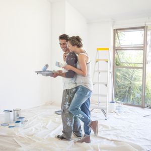 一对年轻的夫妇在油漆溅工作服休息从画房子的内部分享好玩的拥抱。”width=