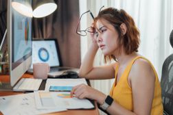 一个坐着的,强调女人看着她的笔记本电脑和图表与她的眼镜”width=