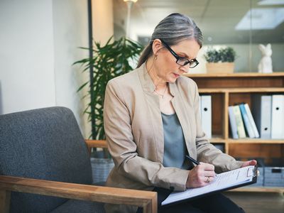 穿着运动上衣和眼镜的女人在办公室的写字板上填写文件＂width=