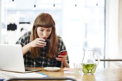 检查她的电话的少妇，当喝在她的膝上型计算机前面的咖啡“width=