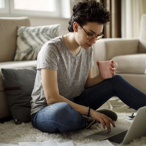 灰色T恤杉的人坐地板，当喝咖啡和做在计算机上的工作时“width=