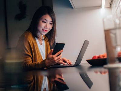 年轻的亚洲女性晚上在家里的厨房里一边用笔记本电脑工作一边看智能手机