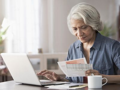 一位年长的妇女正坐在桌子前，将对账单中的信息输入笔记本电脑