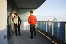 夫妇站在高层阳台上，看着玻璃门“width=