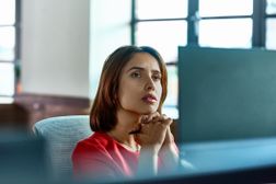 一个女商人坐在电脑屏幕后面，手紧握和凝视，深思熟虑。“width=