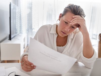 女人看上去很担心在家中拿着文书工作。她正在阅读财务账单或一封有坏消息的信。她看起来很紧张和沮丧。桌子上有一台笔记本电脑。