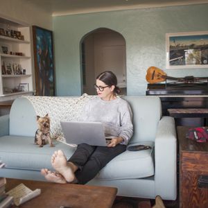 一名戴着眼镜的女人坐在她的创意家中的蓝色沙发上，她的笔记本电脑和小狗“width=