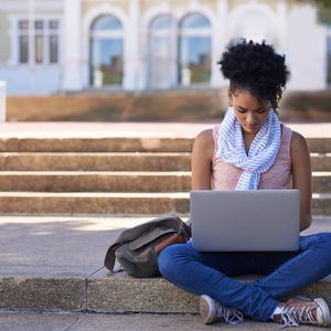 一个大学生坐在外面阴凉的台阶上用笔记本电脑工作。