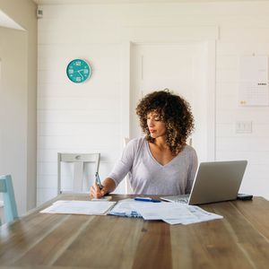 一个女人坐在她的餐厅桌上与膝上型计算机和文书工作做她的税“width=