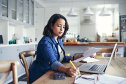 一个女人坐在厨房的桌子旁，用计算器和打开的笔记本电脑计算她的账单。