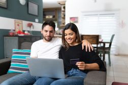 夫妇在笔记本电脑上坐在家里客厅的沙发上购物“width=