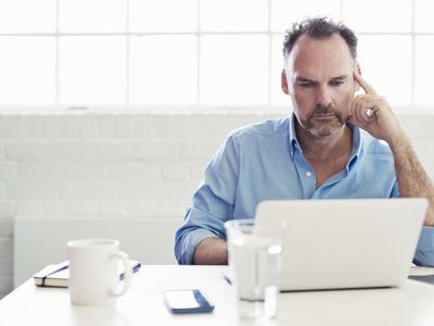 穿着有领衬衫的男人坐在白色的房间里，在电脑前工作＂width=