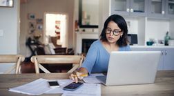 一名妇女坐在厨房桌子旁，用计算器和笔记本电脑计算她的信用卡付款和费用