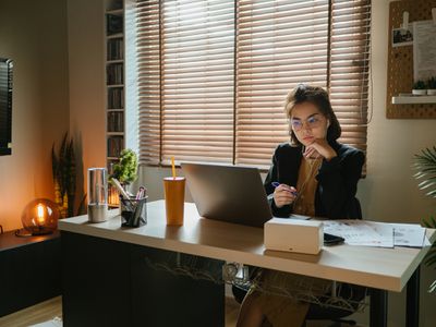 在昏暗的房间里，穿着黑色西装的年轻人坐在桌子前，电脑在她面前