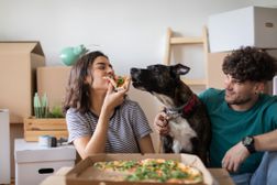 一对年轻的夫妇和他们的狗休息开箱移动箱子在地板上吃披萨的新家”width=