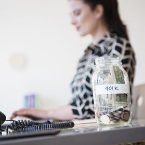 女人在桌子上工作，一罐现金标有401 K坐在桌子上。“width=