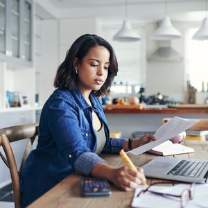 一个女人坐在厨房桌子上，在她的票据与计算器和一个开放的笔记本电脑上工作。“width=
