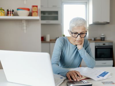 研究在一个计算器的资深妇女在她的厨房用桌附近的膝上型计算机和文件