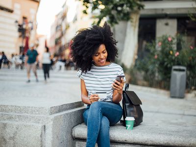 穿着条纹上衣的年轻女子在户外长凳上手持信用卡看着手机。