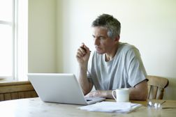 人在他的笔记本电脑坐在厨房的餐桌旁”width=