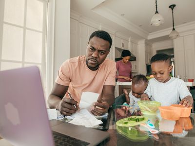 丈夫在孩子玩耍时处理电脑上的财务状况，他的妻子在后台做饭