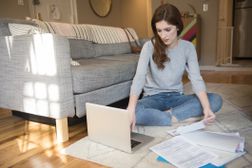 一个年轻的女人坐在地板上的笔记本电脑和文件检查她的401 k。”width=