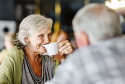 有短的灰色头发的妇女微笑，当从咖啡杯吃一个啜饮时。一个男人用他的回到镜头坐在她身上。他在前景和焦点。“width=