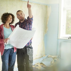 一对年轻夫妇从她的新房子里刮掉墙壁来检查他们的建筑师计划“width=